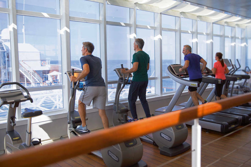 Fitnessraum eines Kreuzfahrtschiffes mit Laufbändern und Ellipsentrainern sowie Panoramafenstern mit Meerblick