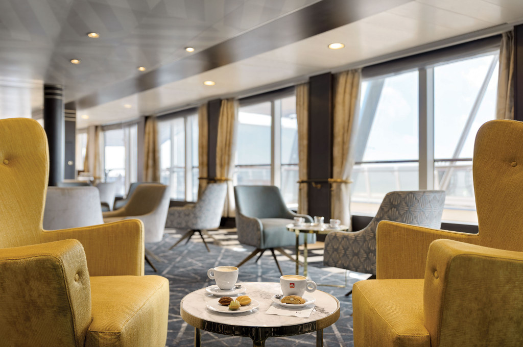 Gemütliche Café-Lounge mit gelben Sesseln und Marmortischen vor Panoramafenstern an Bord eines Hochseekreuzfahrtschiffes.