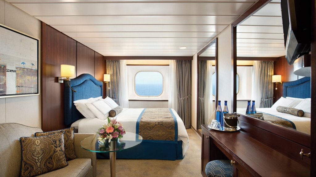 Außenkabine der MS Nautica mit Doppelbett, blauem Polsterkopfteil, Sofa und Meerblick durch Bullauge
