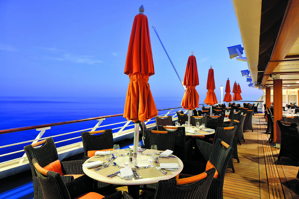 Außenrestaurant mit gedeckten Tischen, orangefarbenen Sonnenschirmen, Korbstühlen und Blick auf das abendliche Meer