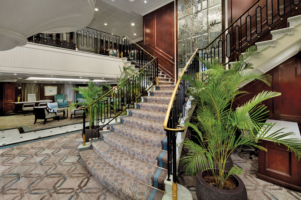 Geschwungene Treppe in der Lobby mit Holzvertäfelung und Palmen auf der Ms Nautica.