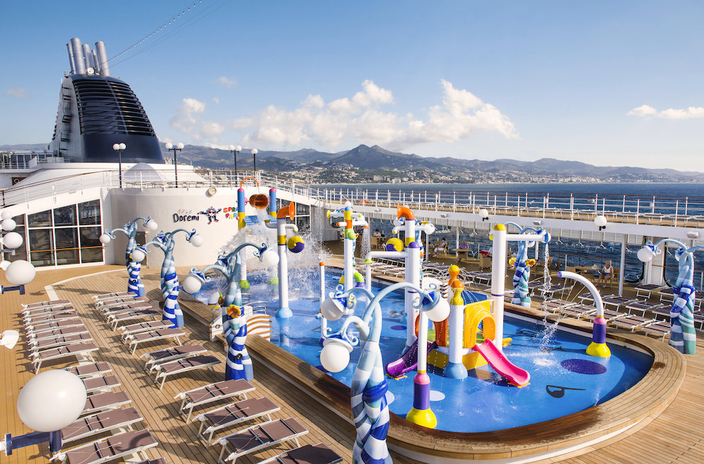 Kinder-Wasserpark auf dem Außendeck eines Kreuzfahrtschiffes mit Wasserspielen und Liegestühlen vor mediterraner Küstenstadt