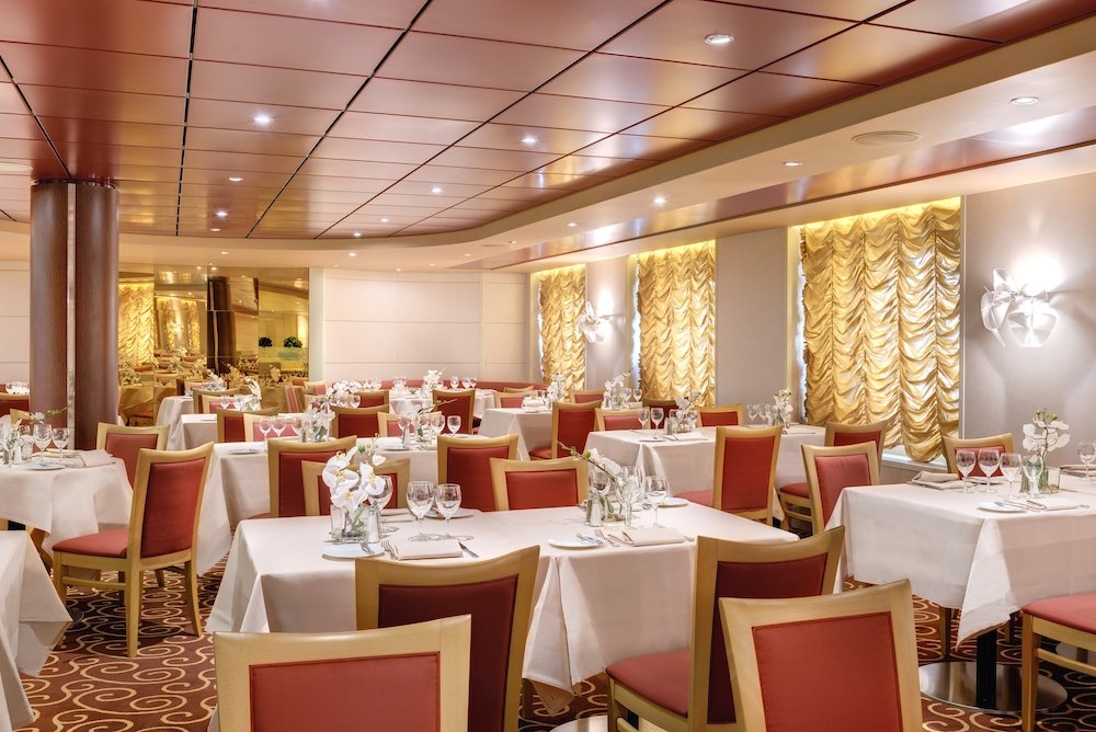 Elegantes Hauptrestaurant mit goldenen Vorhangpaneelen, roten Stühlen, weißen Tischdecken und Orchideenblumen auf der MSC Armonia