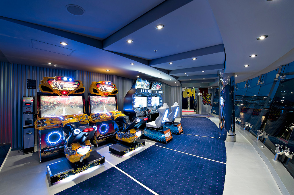 Spielhalle mit Motorrad- und Rennspielsimulationen, bunten Arcade-Automaten und Glaswand mit Meerblick an Bord
