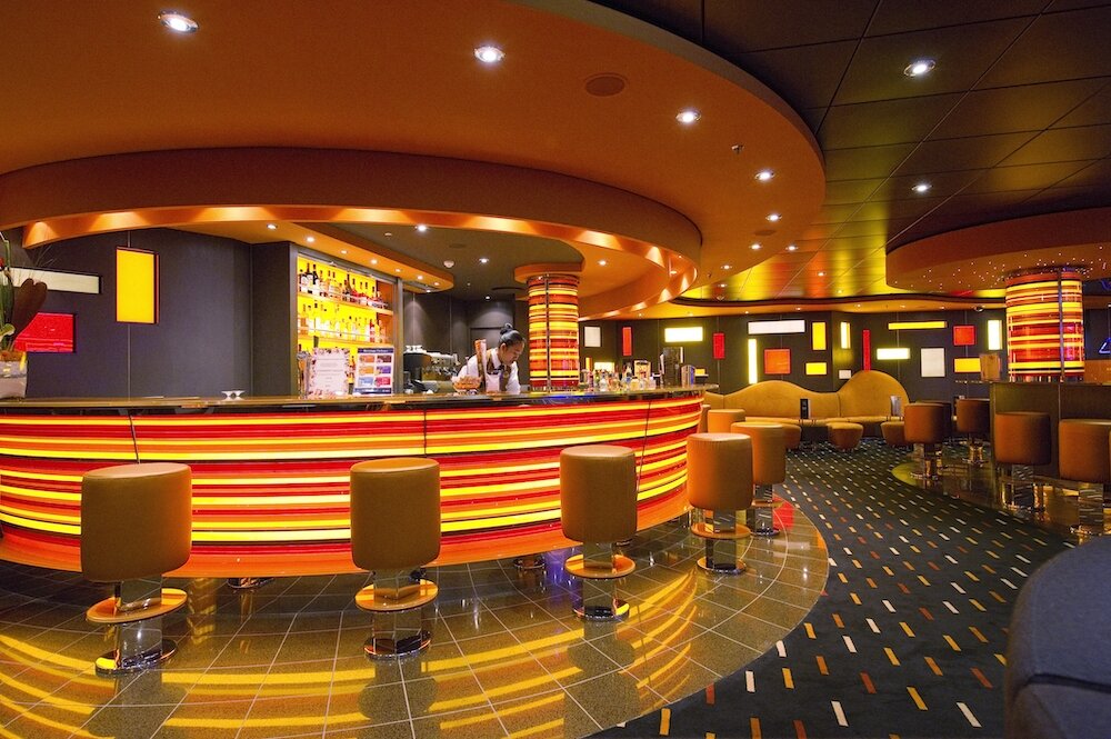 Kreisförmige Bar mit leuchtendem orange-rotem Tresen, Barhockern und Lounge-Sesseln auf einem Kreuzfahrtschiff