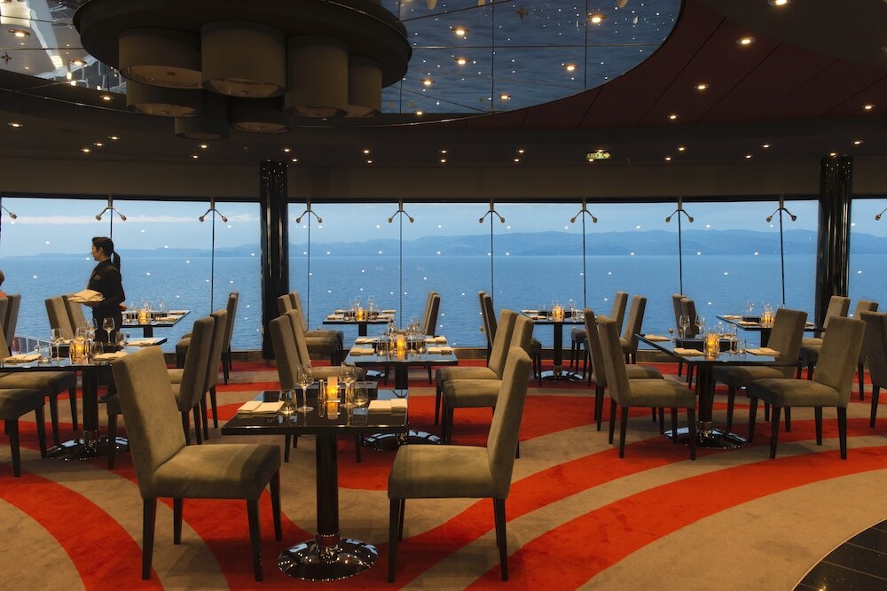 Elegantes Spezialitätenrestaurant mit gestreiftem Teppich, Polsterstühlen, Rundspiegel an der Decke und Panoramafenstern mit Meeresblick bei Abenddämmerung