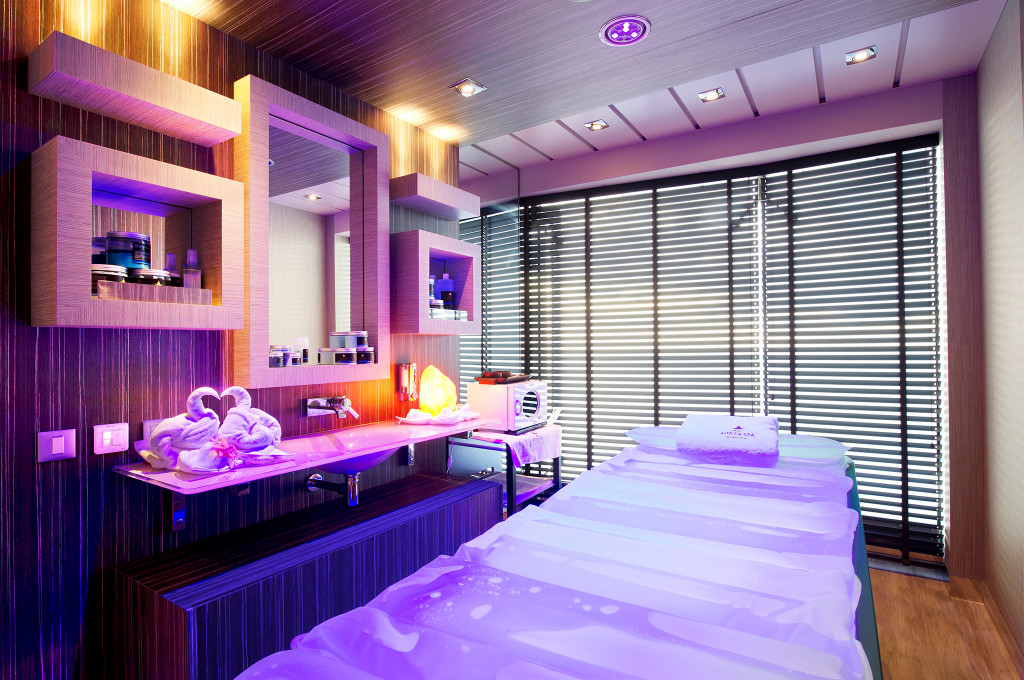 Spa-Behandlungsraum mit Massageliege, violettem Stimmungslicht, Holzwandverkleidung und Produktregalen