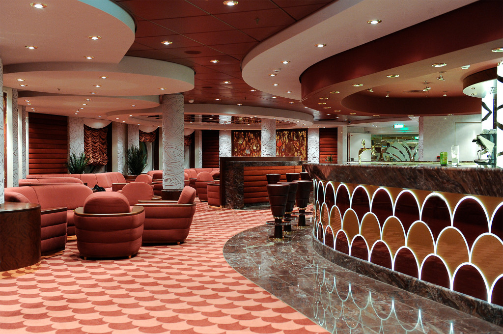 Art-Déco-Bar mit gerundeten rosa Sofas, beleuchtetem Marmortresen mit Muschelrelief-Muster und Rundsäulen auf einem Kreuzfahrtschiff