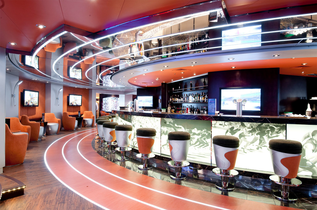 Futuristische Sportsbar mit geschwungenem Bartresen im Rennbahn-Design, orangefarbenen Sesseln, Neonlichtern und Sport-Bildschirmen auf einem Kreuzfahrtschiff