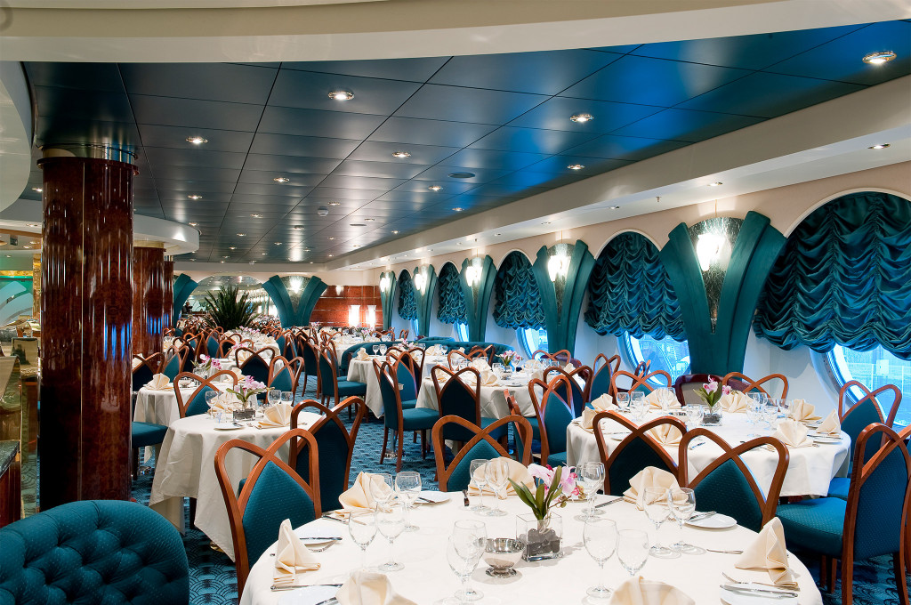 Großes Hauptrestaurant mit runden Tischen, weißen Tischtüchern, Holzstühlen und türkisfarbenen gerafften Vorhängen an Bullaugen auf einem Kreuzfahrtschiff