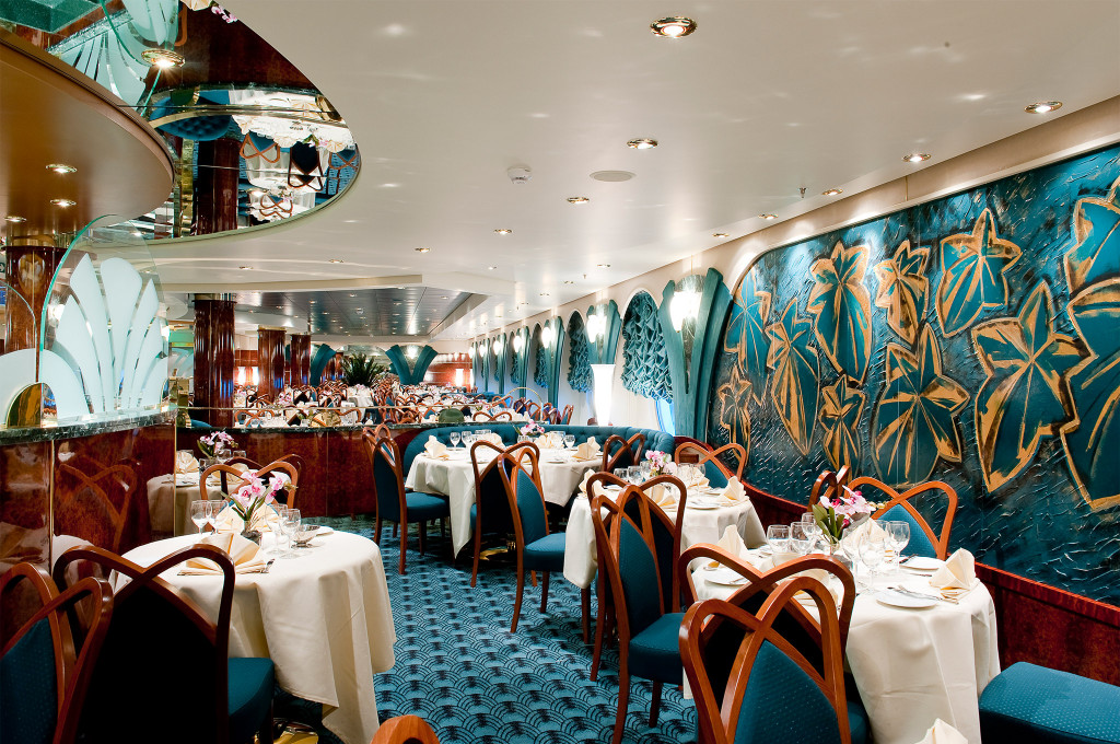 Elegantes Hauptrestaurant mit türkisem Wandrelief mit Goldblatt-Motiv, Holzstühlen mit blauen Polstern und weißen Tischtüchern auf einem Kreuzfahrtschiff