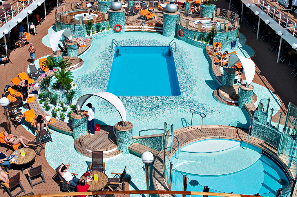 Vogelperspektive des Pooldeck mit türkis-mosaikiertem Pool, Whirlpool, orangefarbenen Liegestühlen und Palmendekor auf einem Kreuzfahrtschiff