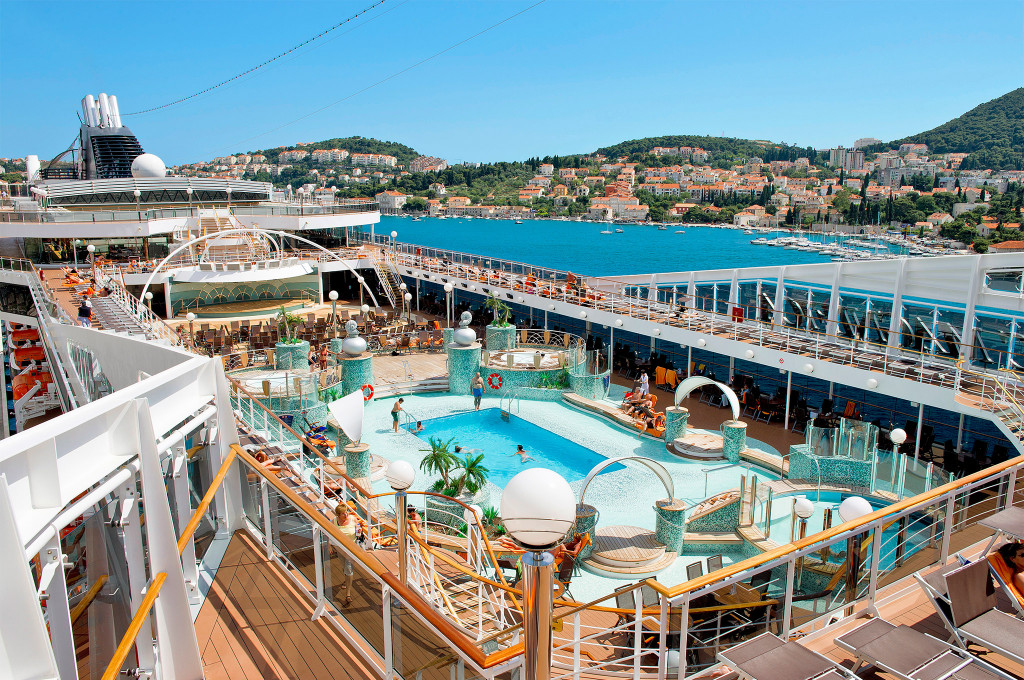 Vogelperspektive des Hauptpooldeck mit zwei Pools, Whirlpools, Sonnenliegen, Palmendekor und Blick auf einen mediterranen Hafen