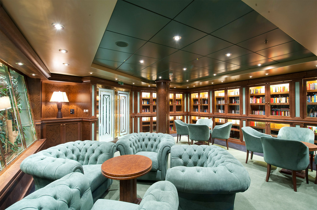 Elegante Bibliothekslounge mit mintgrünen Chesterfield-Sofas, beleuchteten Mahagoni-Bücherregalen und Tischlampe auf einem Kreuzfahrtschiff
