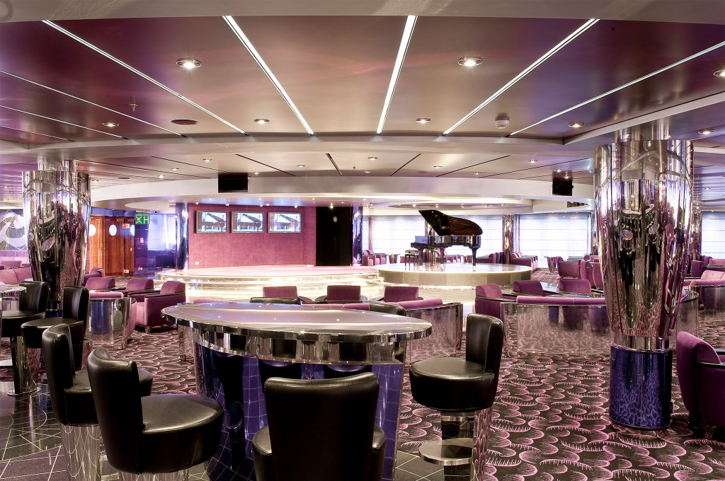 Weitläufige lila Lounge mit runden Chromstehtischen, schwarzen Drehstühlen, Bühne mit Flügel und Panoramafenstern auf einem Kreuzfahrtschiff
