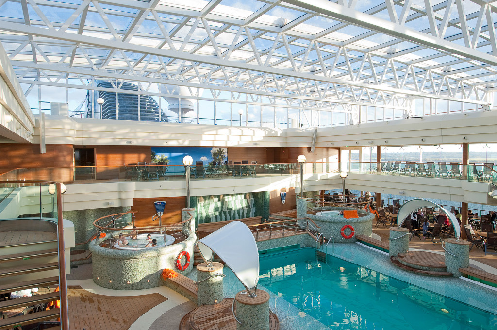 Überdachter Innenpool mit Glasdach, zwei Whirlpools, Liegestühlen auf Galerie-Ebene und Blick auf die Aufbauten des Kreuzfahrtschiffs
