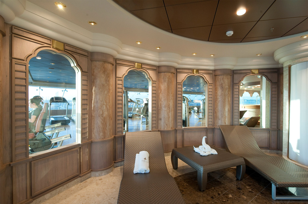 Spa-Ruheraum mit Rattanliegen, gefalteten Handtüchern und Bogenfenstern zum angrenzenden Fitnessstudio auf einem Kreuzfahrtschiff