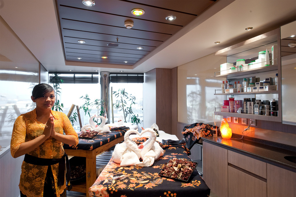 Spa-Behandlungsraum mit zwei Massageliegen mit Handtuchschwänen, Pflege-Produkten und balinesischer Therapeutin auf einem Kreuzfahrtschiff