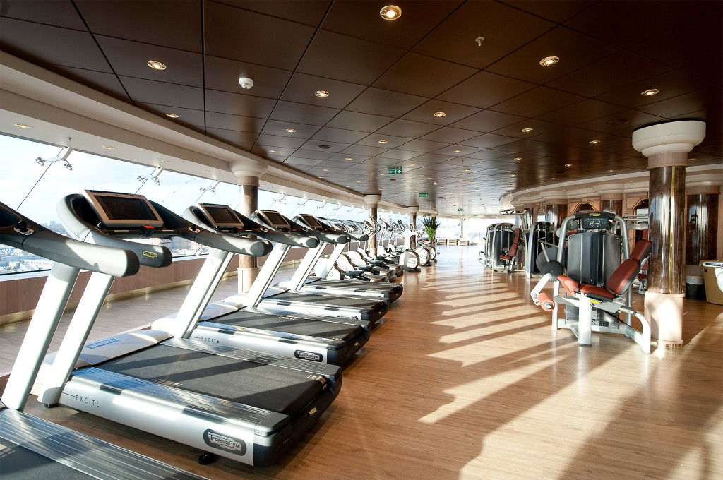 Großes Fitnessstudio mit einer Reihe Laufbänder, Kraftgeräten, Holzboden und großen Fenstern auf einem Kreuzfahrtschiff