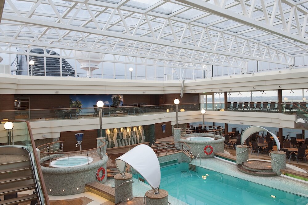 Überdachter Poolbereich eines Kreuzfahrtschiffes mit Glasdach, Pool, Whirlpools und umlaufender Terrasse