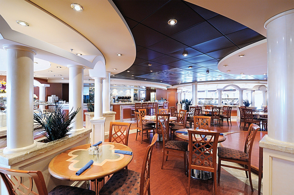 Klassisches Hauptrestaurant auf einem Kreuzfahrtschiff mit weißen Säulen, runden Tischen mit blauen Servietten und Buffettheke im Hintergrund