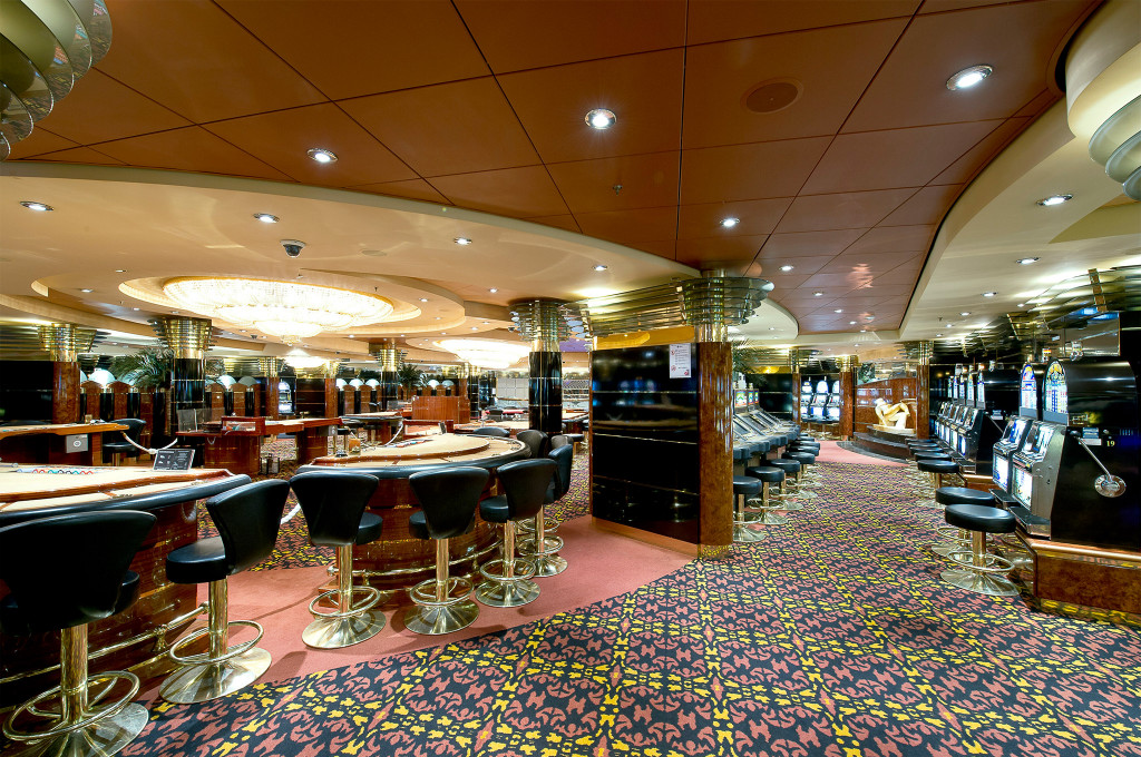 Großes Casino auf einem Kreuzfahrtschiff mit Spieltischen und Spielautomaten in goldfarbenem Interieur mit gemustertem Teppich