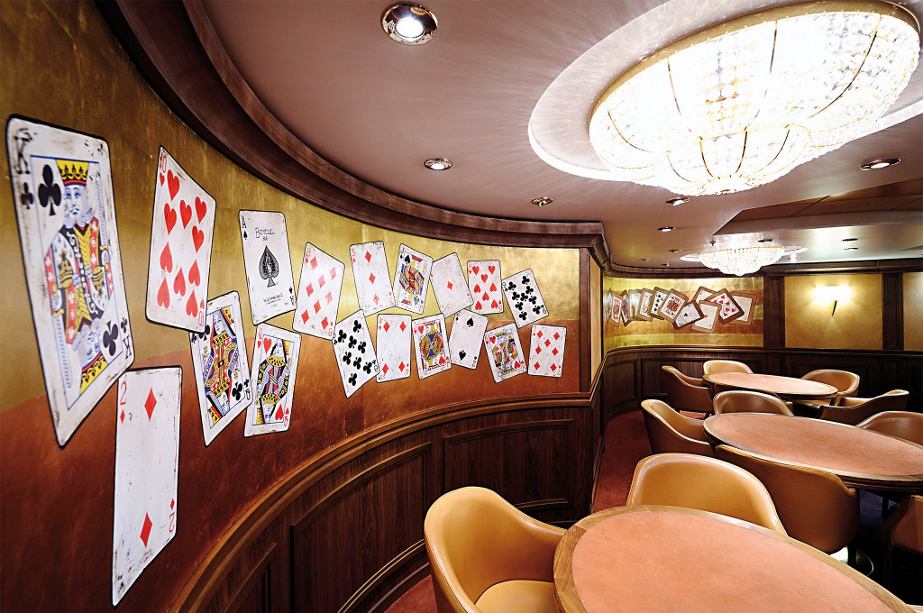 Kartenspielzimmer auf einem Kreuzfahrtschiff mit vergoldeten Wänden voller Spielkartenmotive, Filztischen und Kristallkronleuchter