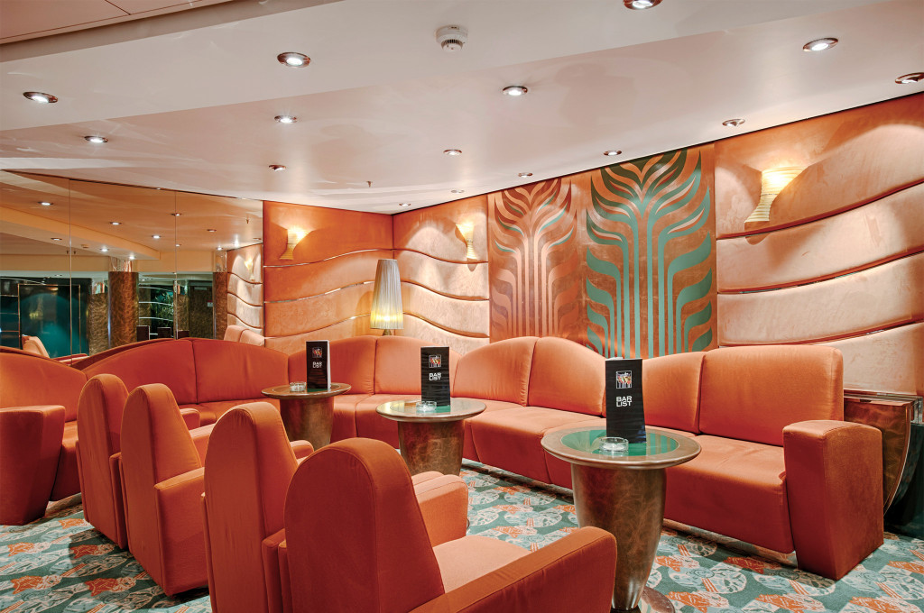 Lounge auf einem Kreuzfahrtschiff mit orangefarbenen Samtsofas, geschwungenen Wandpaneelen mit Art-Déco-Motiven und Beistelltischen