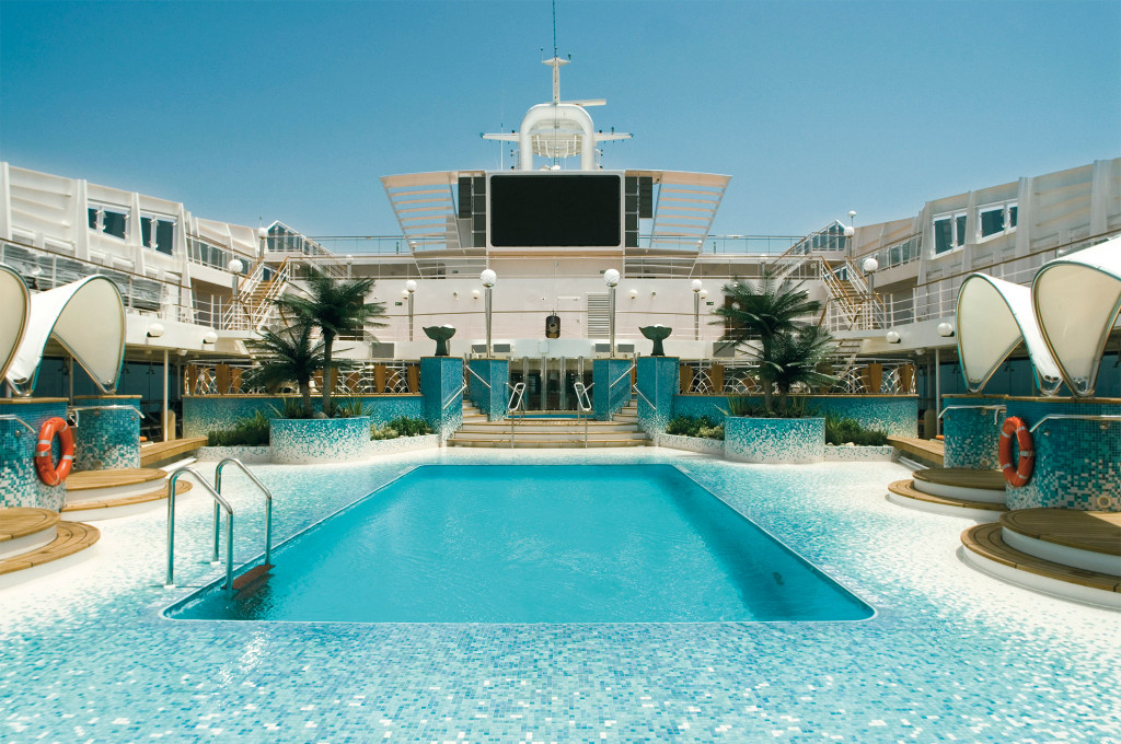 Pooldeck auf einem Kreuzfahrtschiff mit rechteckigem Pool, türkisen Mosaikfliesen, Palmen und großem Bildschirm im Hintergrund