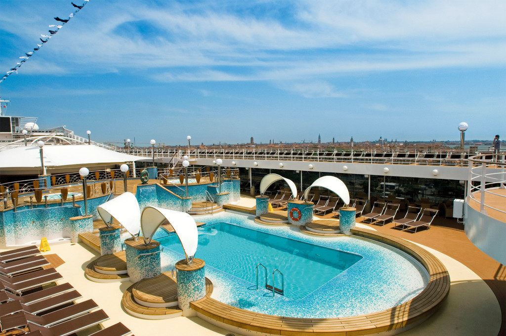 Pooldeck eines Kreuzfahrtschiffes mit Pool, Mosaikfliesen und geschwungenen Sonnensegeln vor der Skyline von Venedig