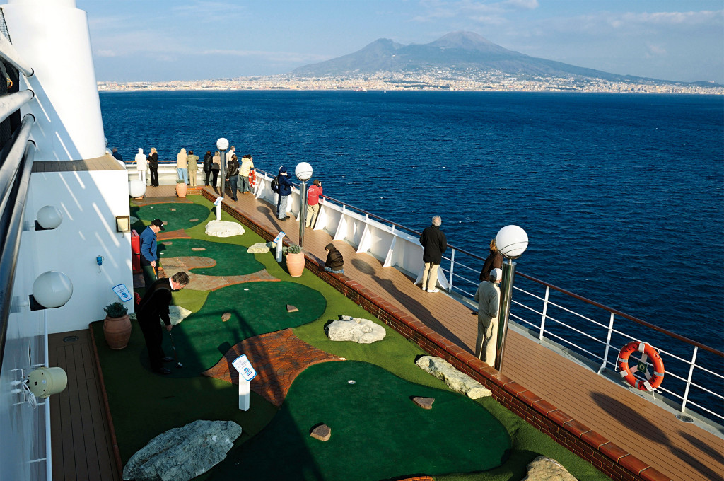 Gäste spielen Minigolf auf dem Außendeck eines Kreuzfahrtschiffes mit dem Vesuv und der Küste von Neapel im Hintergrund