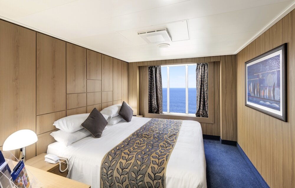 Außenkabine mit Doppelbett, Holzvertäfelung und Panoramafenster mit Meerblick auf einem Kreuzfahrtschiff