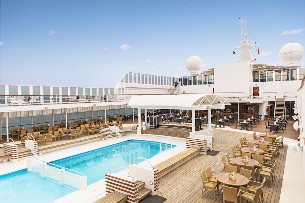 Elegantes Pooldeck eines Kreuzfahrtschiffes mit Pool, Liegestühlen, Außenterrasse und Bar unter blauem Himmel