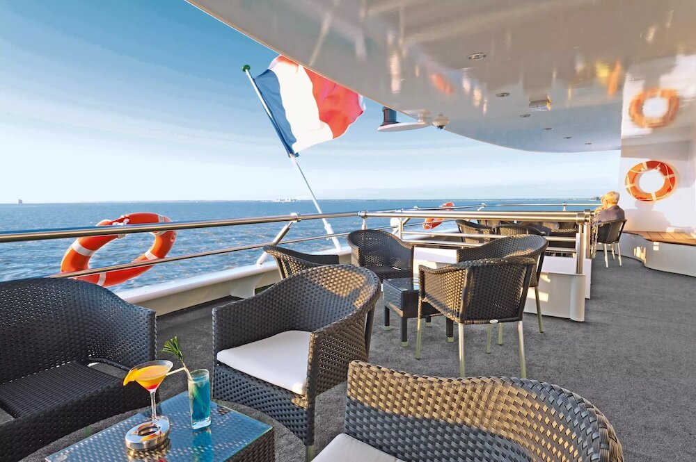 Sonnige Außenterrasse der MS Cyrano de Bergerac von nicko cruises mit dunklen Rattanstühlen und Glastischen, an denen ein Cocktail und ein blaues Getränk stehen. Die französische Trikolore weht am Heck, im Hintergrund glitzert das Meer, orangefarbene Rettungsringe hängen an der Reling.