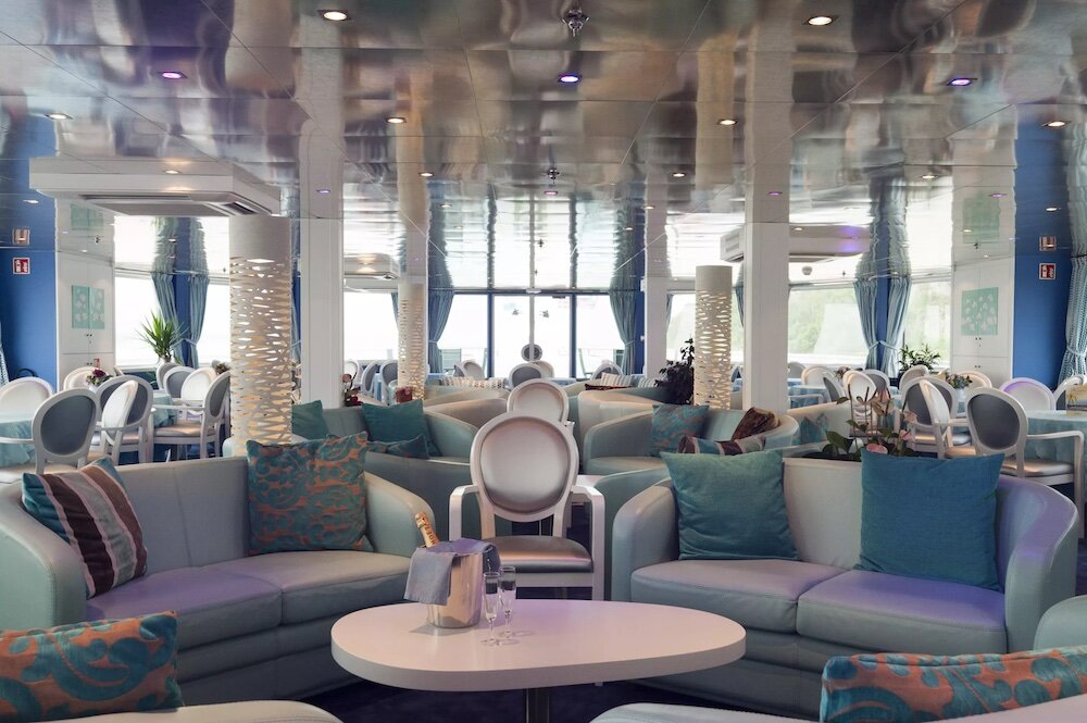 Heller Salon eines Flusskreuzfahrtschiffes mit Sofas, Sesseln, ovalem Couchtisch mit Champagnerkübel, dekorativen Säulen und Panoramafenstern