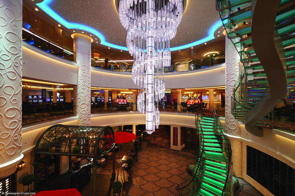 Mehrstöckiges Atrium Ocean Place mit Kristallkronleuchter und grün beleuchteter Glastreppe an Bord der Norwegian Getaway.