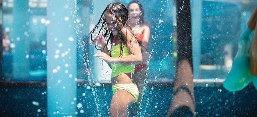 Mädchen läuft lachend durch Wasserfontänen im Kinderwasserpark auf dem Deck eines Hochseekreuzfahrtschiffes
