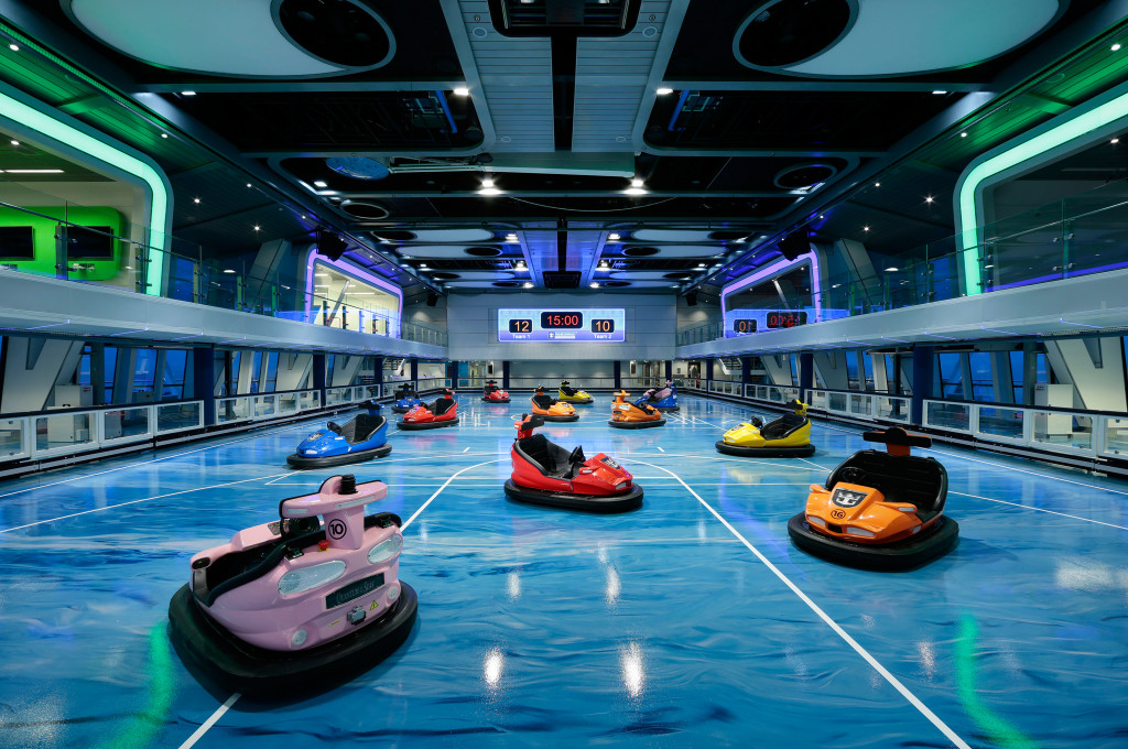 Indoor-Sporthalle mit bunten Autoscootern auf blauer Fahrbahn und Anzeigetafel an Bord eines Kreuzfahrtschiffs.