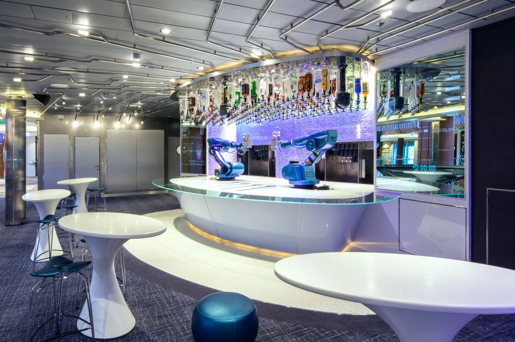 Futuristische Bar mit zwei Roboterarmen als Barkeeper, Flaschenwand und weißen Stehtischen an Bord eines Kreuzfahrtschiffs.