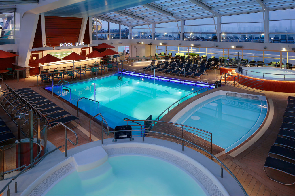 Überdachter Innenpool mit Whirlpools, Liegestühlen und Poolbar unter einem Glasdach an Bord eines Kreuzfahrtschiffs.