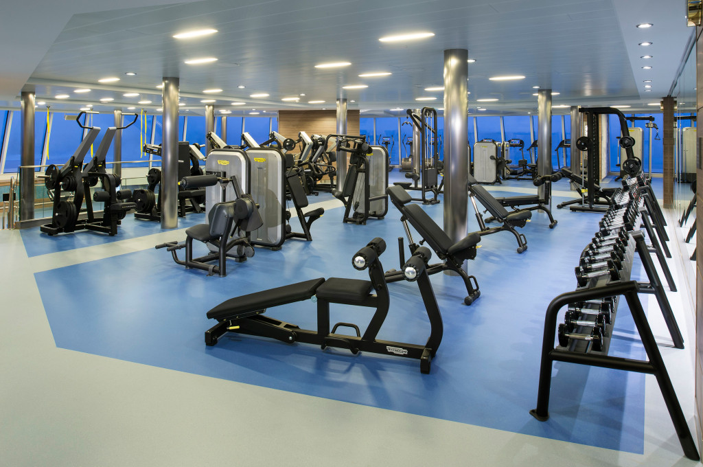 Bordfitnesscenter mit Kraftgeräten, Hantelablage und Bänken vor Panoramafenstern an Bord eines Kreuzfahrtschiffs.