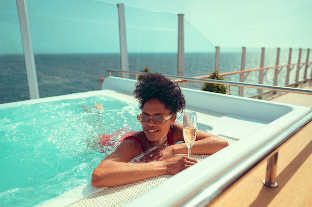 Frau im Außenwhirlpool eines Kreuzfahrtschiffes mit Champagnerglas und Blick auf das offene Meer
