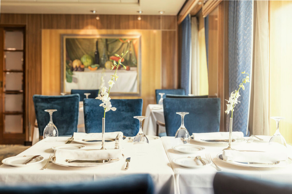 Elegantes Restaurant der Queen Mary 2 mit blauen Samtsesseln, weiß gedeckten Tischen und Orchideenvasen