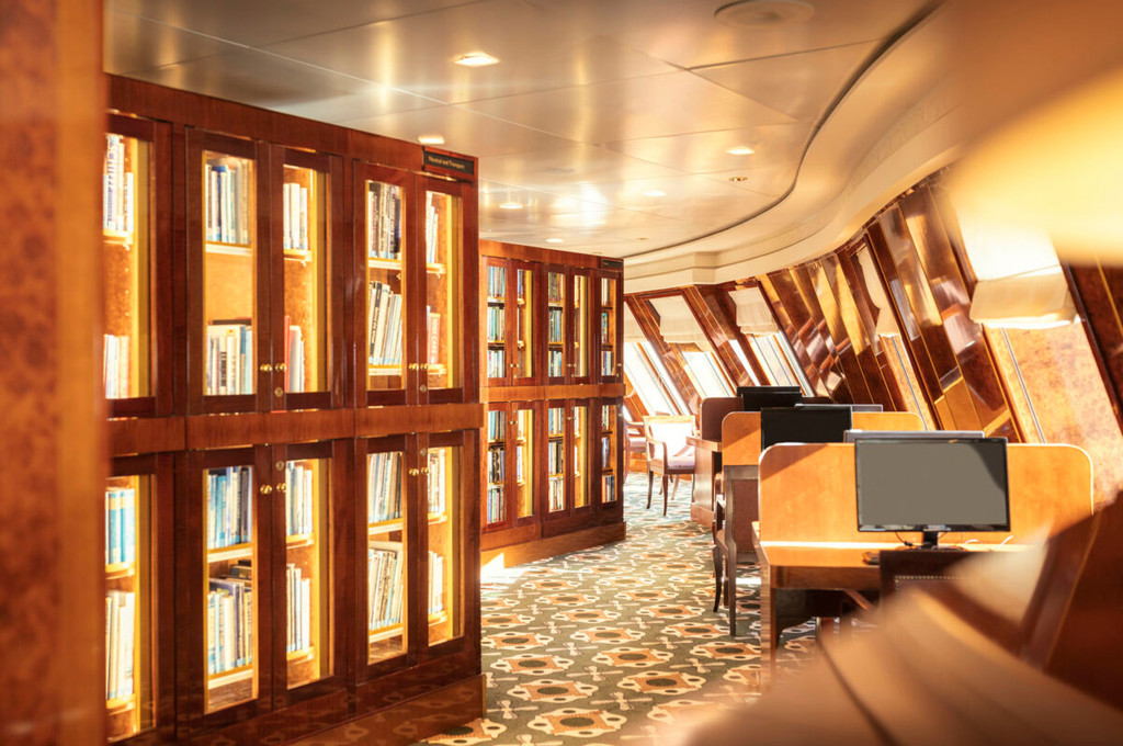 Bibliothek der Queen Mary 2 mit beleuchteten Bücherregalen, Computerarbeitsplätzen und schrägen Schiffsfenstern