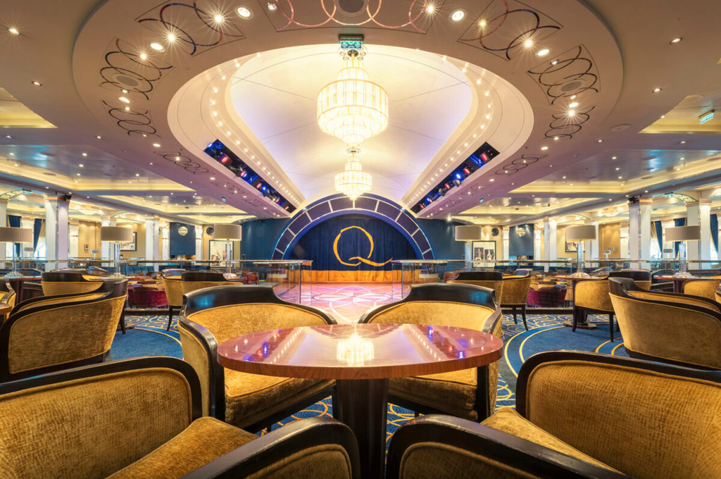 Ballsaal-Lounge der Queen Mary 2 mit goldenen Sesseln, Rundbühne mit Q-Logo und prächtigem Kronleuchter