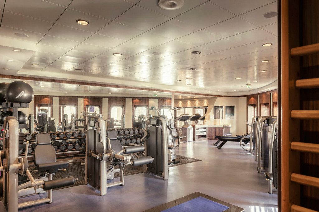 Weitläufiger Fitnessraum der Queen Mary 2 mit Kraftgeräten, Hantelregal und Spiegelwand