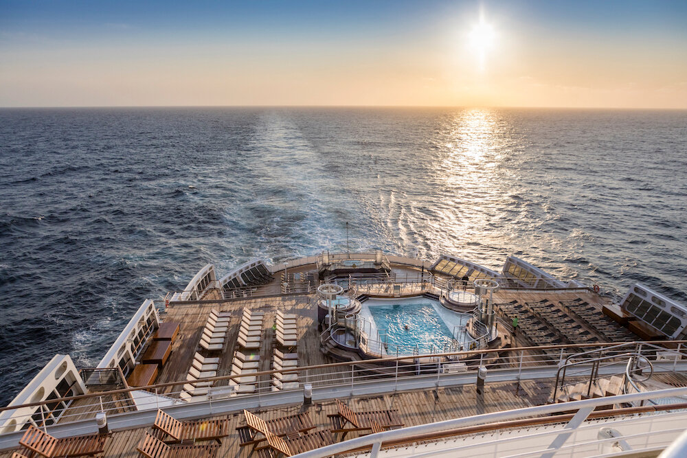 Pooldeck der Queen Mary 2 bei Sonnenuntergang mit Liegestühlen, Heckwelle und weitem Meerblick am Horizont