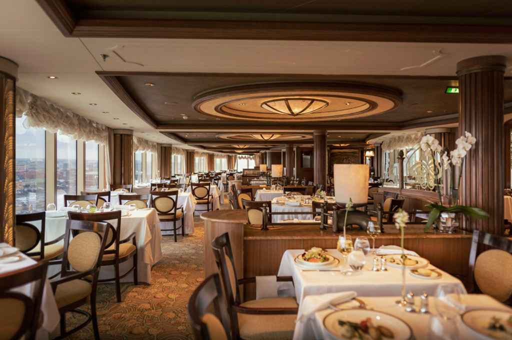 Elegantes Restaurant der Queen Victoria mit Panoramafenstern, weiß gedeckten Tischen und rundem Art-déco-Deckenornament