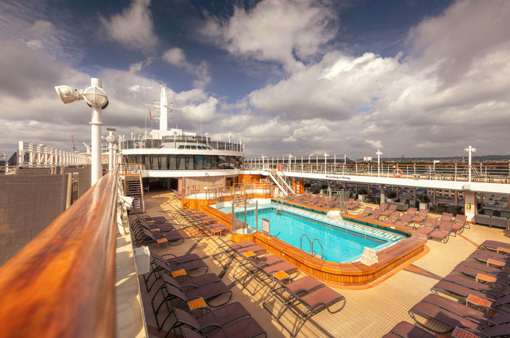 Sonnendeck der Queen Victoria mit Außenpool, Teakholzumrahmung und Liegestühlen unter bewölktem Himmel
