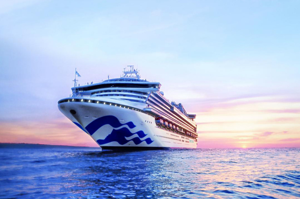 Großes Kreuzfahrtschiff mit blauem Wellensymbol am Bug fährt bei pastellfarbenem Sonnenuntergang auf ruhiger See.